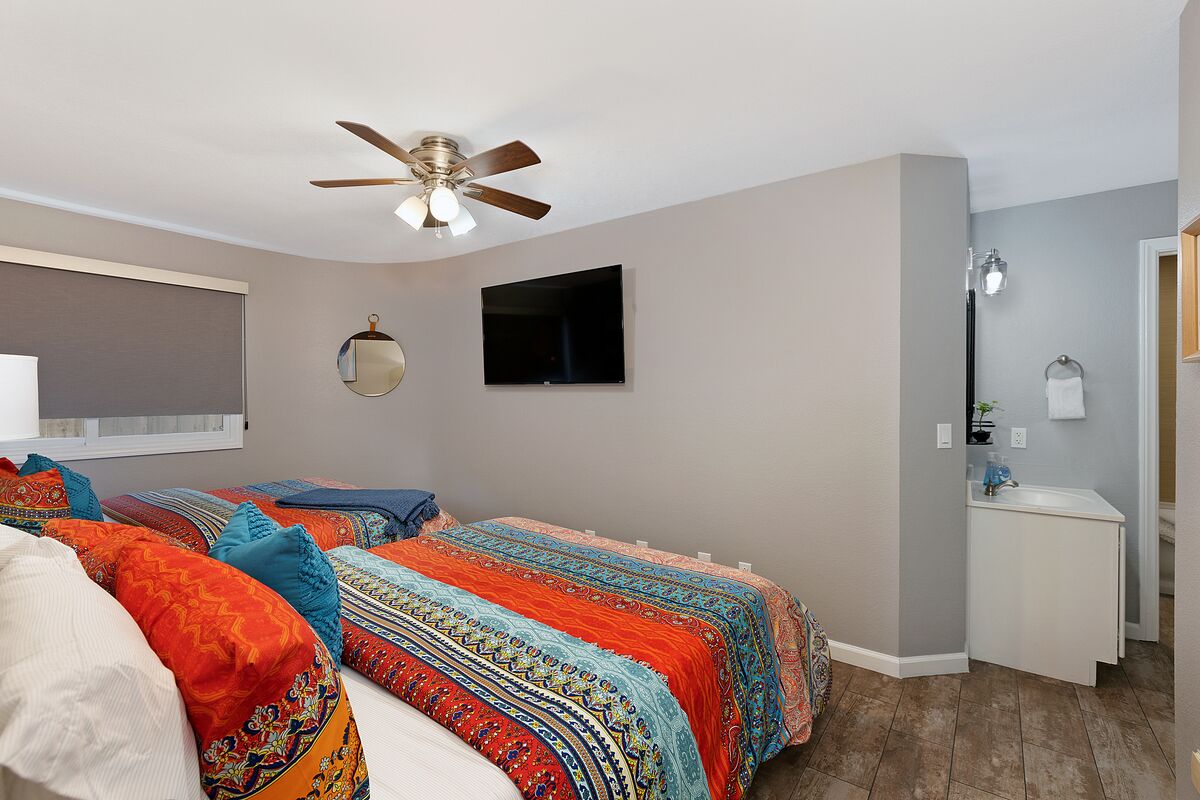 A bedroom with two beds, a ceiling fan, a TV, a mirror, and a bathroom with a sink and towel rack.