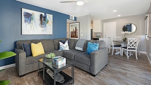 A modern living room with a gray sectional sofa, a glass coffee table, and a dining area with white chairs and a table.