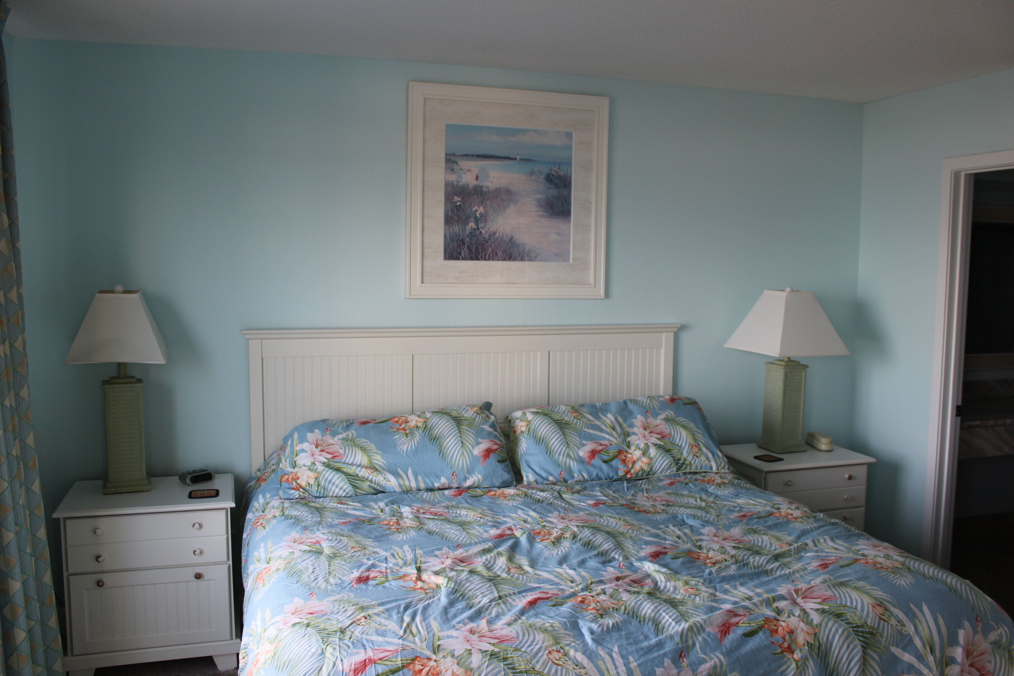 A bedroom with a bed, two nightstands, a framed picture on the wall, and a door to the right.
