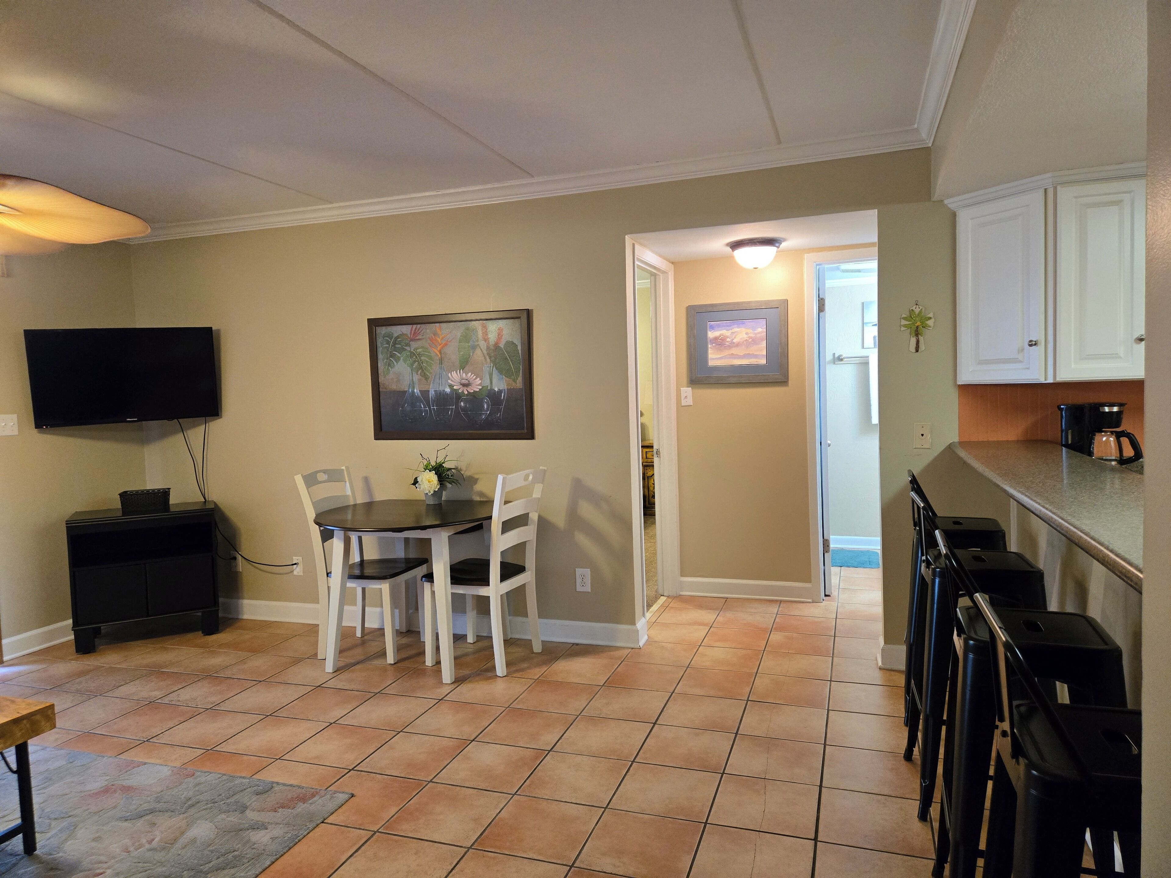 A dining area with a table and chairs, a television mounted on the wall, and a kitchen area with cabinets and a countertop.