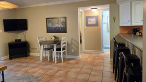 A dining area with a table and chairs, a television mounted on the wall, and a kitchen area with cabinets and a countertop.