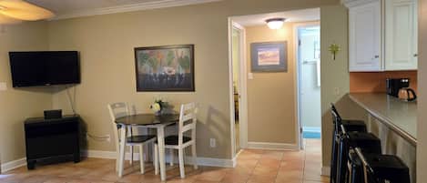 A dining area with a table and chairs, a television mounted on the wall, and a kitchen area with cabinets and a countertop.