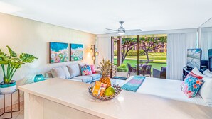 A modern hotel room with a large window, a ceiling fan, a dining area with outdoor seating, and a fruit basket on the counter.