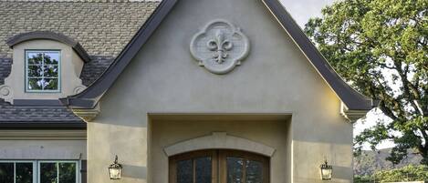 A house with a symmetrical facade, a central entrance with a wooden door, and a decorative emblem above the door. There are two potted plants on either side of the entrance, and a gravel driveway leading to the house.