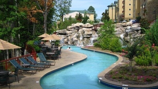 A swimming pool with a waterfall feature, surrounded by lounge chairs and tables, with a stone wall and greenery in the background. - Riverstone 2BR - River & Mountain Views / 5 min to Dollywood & Ripken (Pigeon Forge)