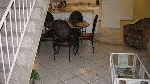 A dining area with a round table and four chairs, a kitchen in the background, a staircase, and a patterned sofa. - 3rd Floor Incredible Location-Wifi-2 BR/2 BA Loft Sleeps 8 (Orange Beach)