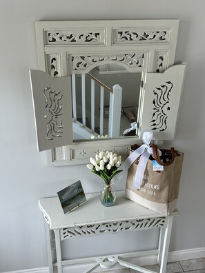 Um console branco com um espelho, um vaso de flores brancas, um quadro emoldurado e um saco de papel castanho com texto.