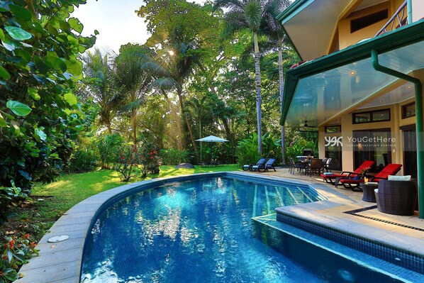 A swimming pool with a glass-enclosed patio area, surrounded by lush greenery and a well-maintained lawn.