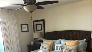 Un dormitorio con cama, ventilador de techo, mesita de noche, lámpara y ventana con cortinas.