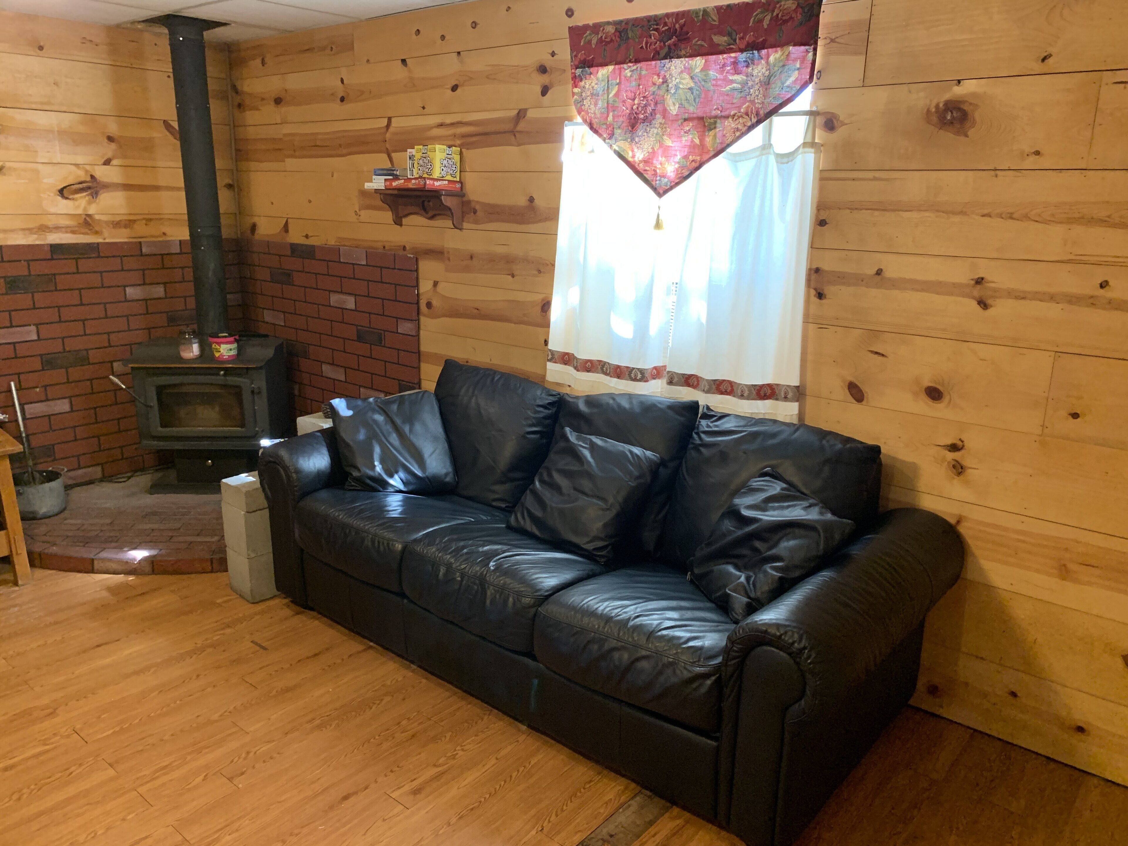 A cozy room with a black leather sofa, a brick fireplace, wooden walls, and a window with curtains.