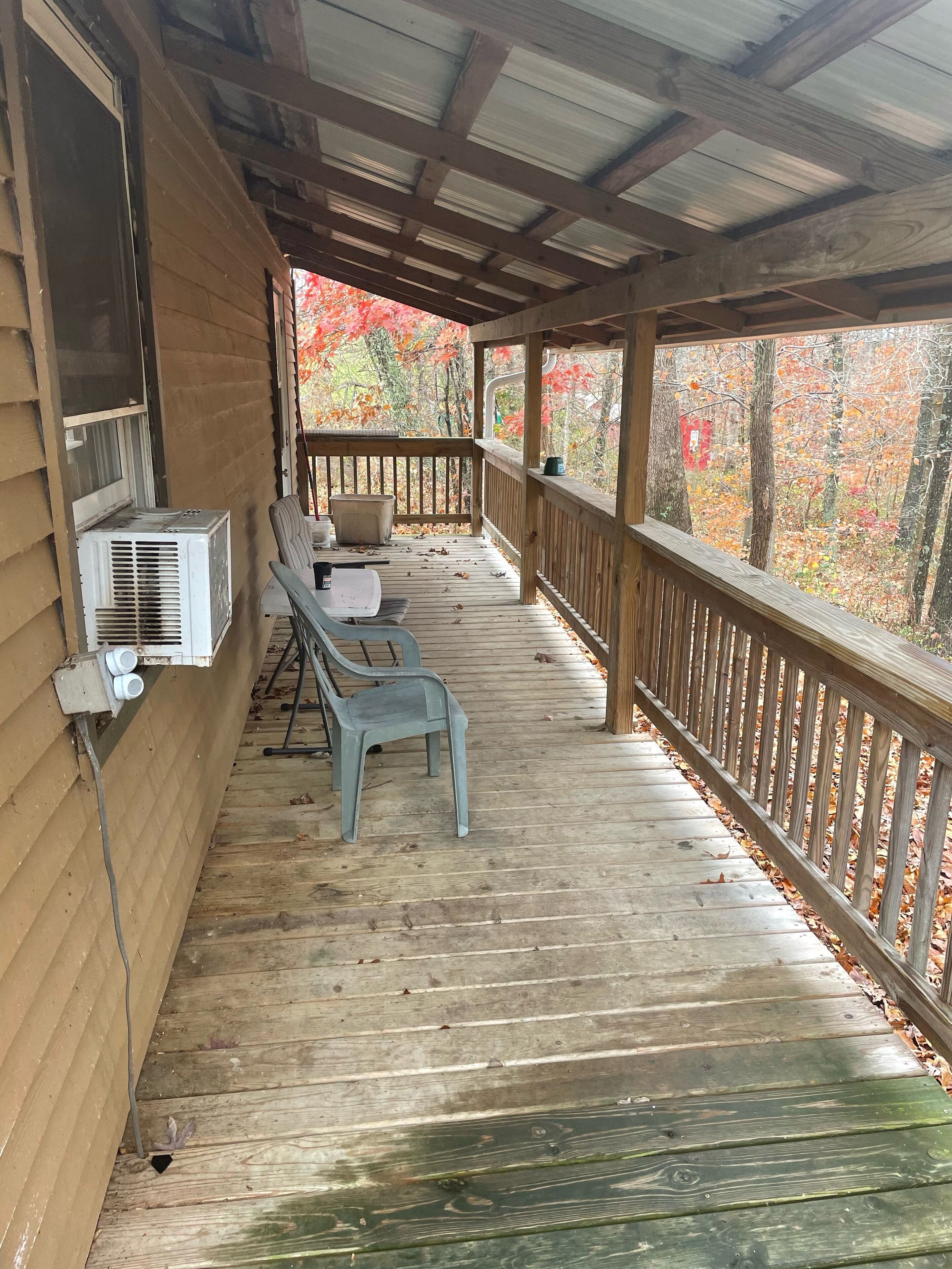 A covered porch with a wooden railing, a window with an air conditioning unit, and a chair.