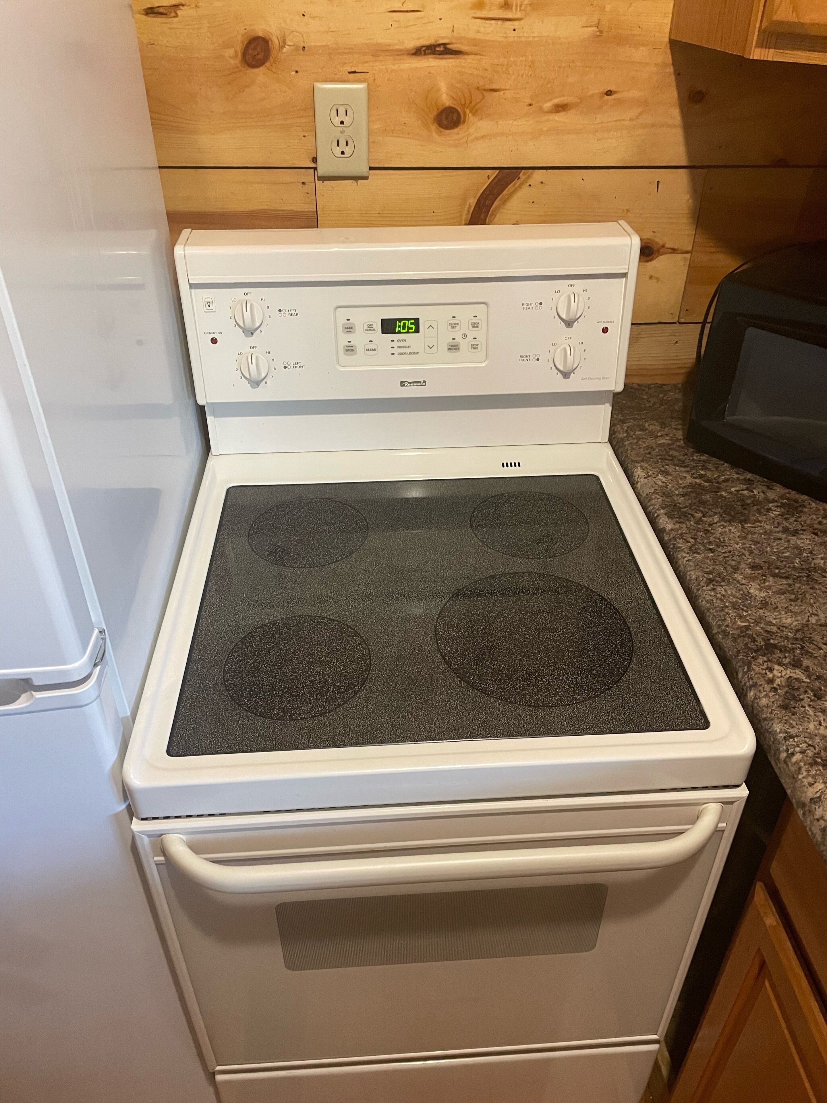 Um fogão elétrico branco com visor de relógio digital, painel de controle com vários botões e porta de forno com ventilação.