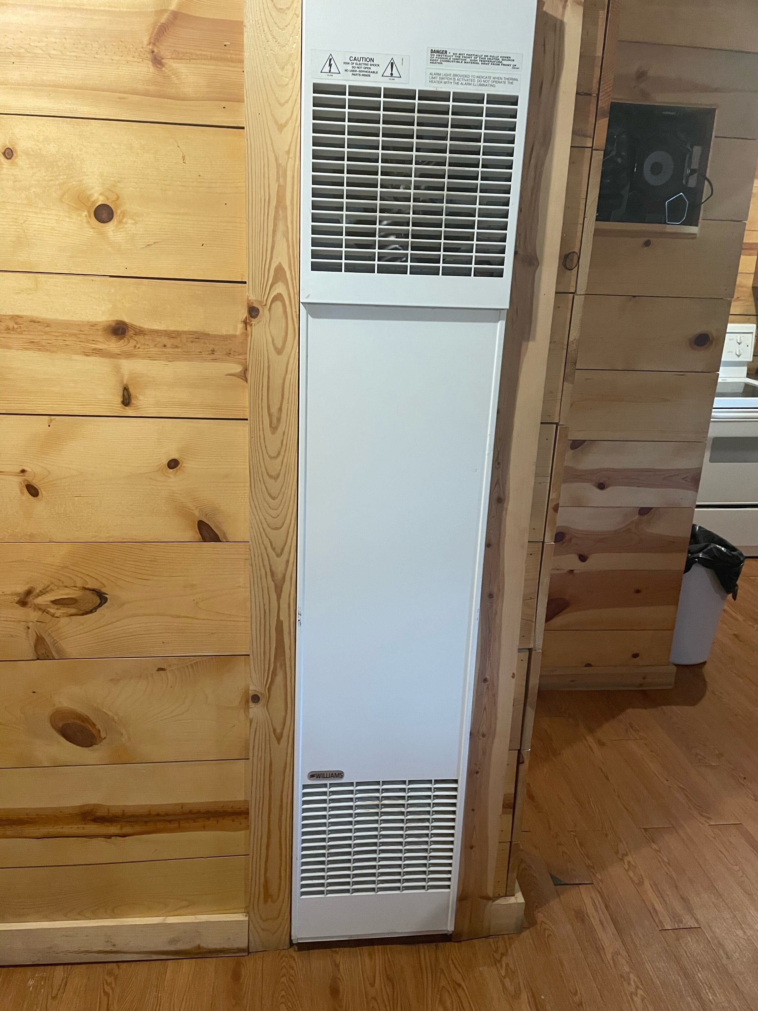 Un equipo de aire acondicionado blanco con etiqueta de advertencia y una rejilla de ventilación en la parte inferior, apoyado contra una pared de madera con vetas y nudos visibles.