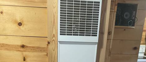 Un equipo de aire acondicionado blanco con etiqueta de advertencia y una rejilla de ventilación en la parte inferior, apoyado contra una pared de madera con vetas y nudos visibles.