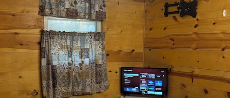 Une chambre à coucher avec un lit, un ventilateur de plafond, une fenêtre avec des rideaux à motifs et un téléviseur posé sur une petite table.