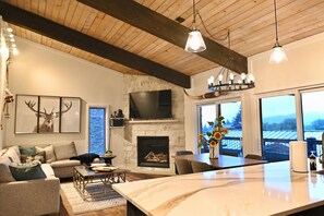A modern living room with a fireplace, a large TV mounted on a stone wall, and a dining area with a marble island. - Newly Remodeled Ski-In, Ski-Out To Lift About 100 Yards From Unit. (Snowmass Village)