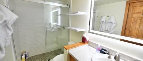 A bathroom with a glass shower enclosure, a toilet, a sink with a mirror, and wooden cabinets.