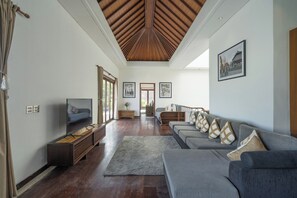 A spacious living area with a high wooden ceiling, a grey sofa set, a TV, and framed pictures on the walls.