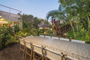 Un espace extérieur abrité, aménagé pour les repas, avec une table et des chaises carrelées, entouré d’une végétation luxuriante et offrant une vue sur un bâtiment en arrière-plan.
