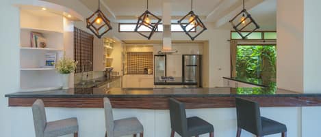 A modern kitchen with a central island, bar stools, pendant lighting, and a view of the greenery outside.
