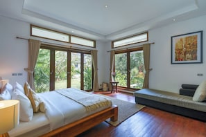 A spacious bedroom with a large bed, a sofa, and a view of a garden through large windows.