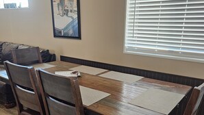 A dining area with a wooden table and chairs, a framed picture on the wall, and a window with blinds. - Movie House (Thompson)