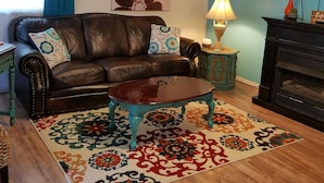 A living room with a dark brown leather sofa, a wooden coffee table, a patterned rug, a ceiling fan, a wall-mounted TV, and a fireplace.