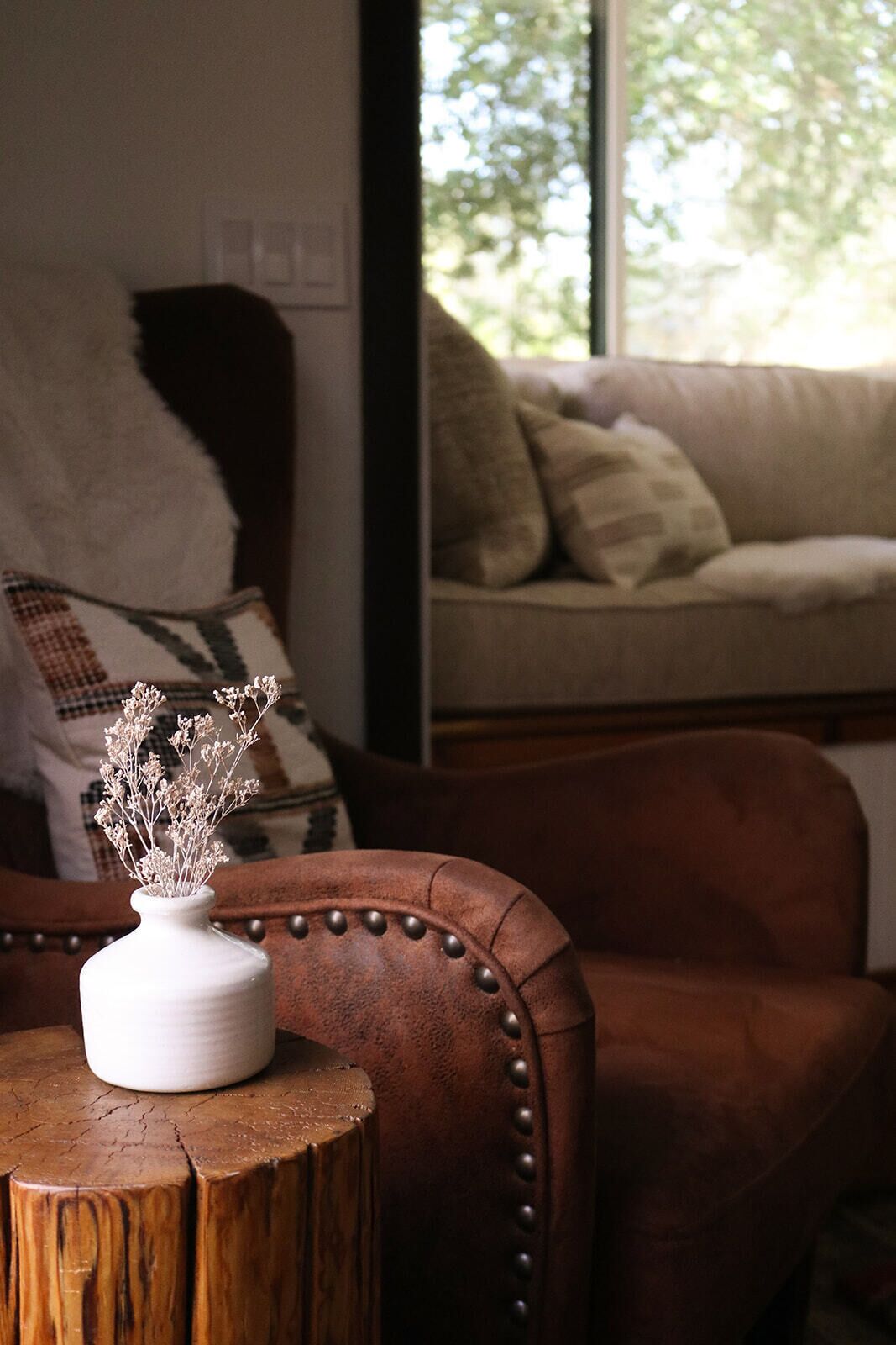 A rustic wooden table with a white vase containing dried flowers, positioned in front of a leather armchair. A sofa with patterned cushions is visible in the background.