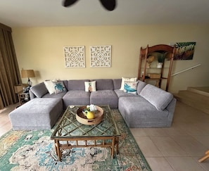A living room with a grey sectional sofa, a glass coffee table with a woven basket, patterned rug, and decorative wall art.