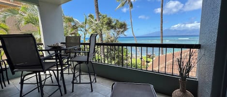 A balcony with a view of the ocean, palm trees, and a dining area with chairs and a table.