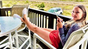 Eine Person sitzt auf einem Liegestuhl mit einem Tisch und blickt auf einen Strand und eine Brücke.