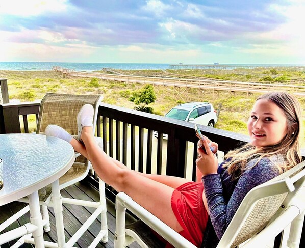 A person sitting on a deck chair with a table, overlooking a beach and a bridge.
