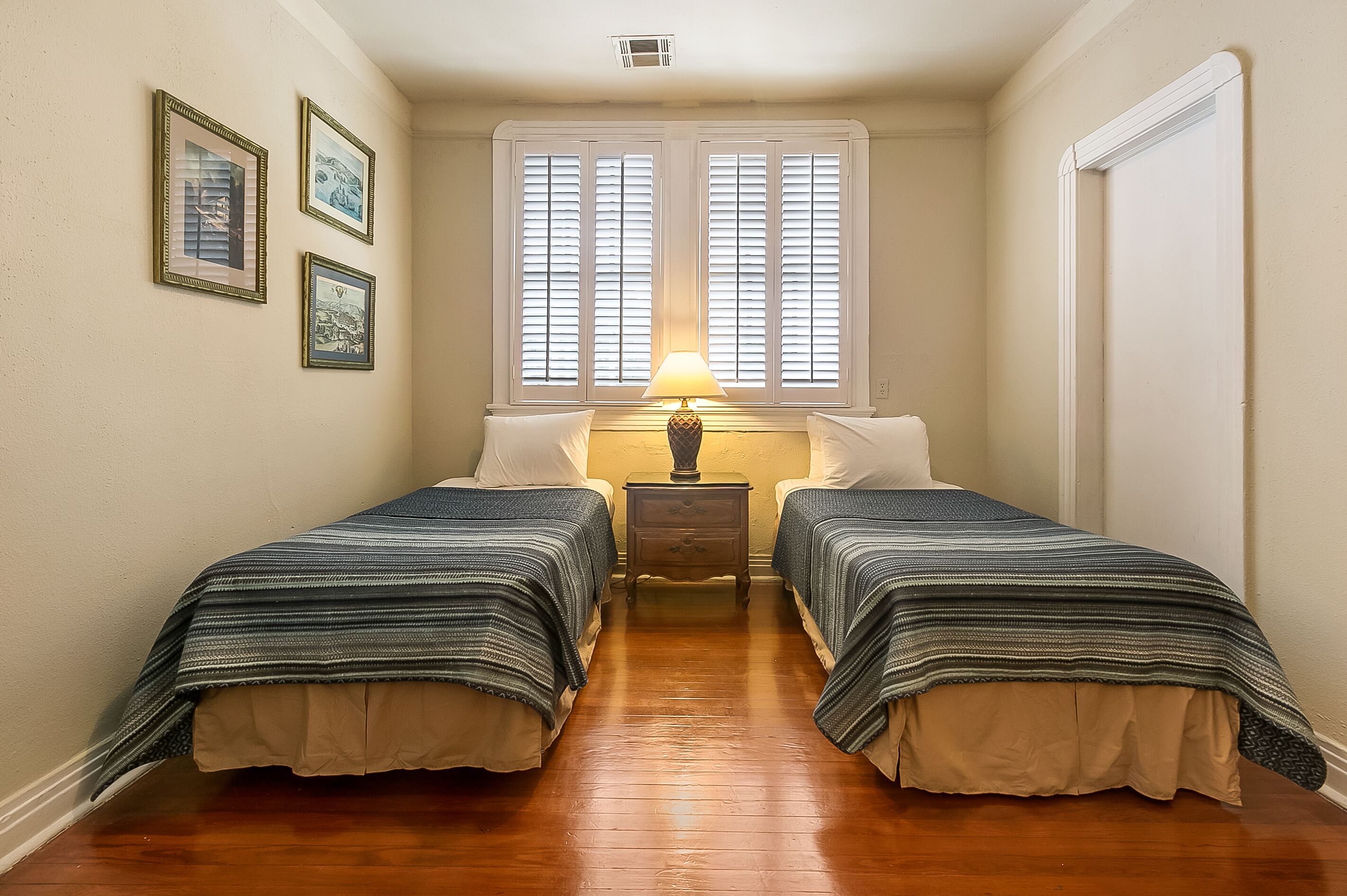 A narrow room with two beds, a nightstand with a lamp, and three framed pictures on the wall.