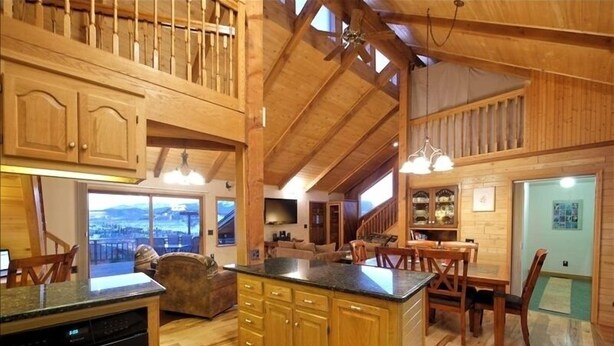 Una cocina amplia con gabinetes de madera, encimera de granito y una isla grande. Hay un comedor con sillas y mesa de madera, y vistas a un paisaje urbano a través de amplios ventanales.