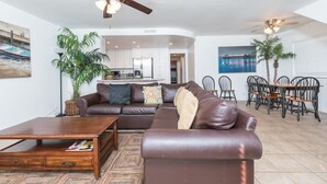 A living room with a brown leather sofa, a wooden coffee table, a dining area with a wooden table and chairs, a kitchen in the background, and a ceiling fan.
