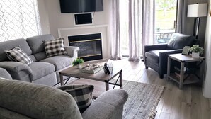 Ein modernes Wohnzimmer mit einem grauen Sofagarnitur, einem Couchtisch, einem Kamin und einem an der Wand befestigten Fernseher.
