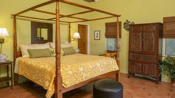 A four-poster bed with a floral bedspread, a wooden nightstand with a lamp, a potted plant, and a wooden cabinet with intricate carvings.