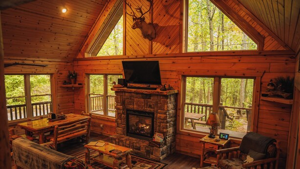 A cabin interior with a stone fireplace, wooden furniture, and a deer head mounted on the wall.