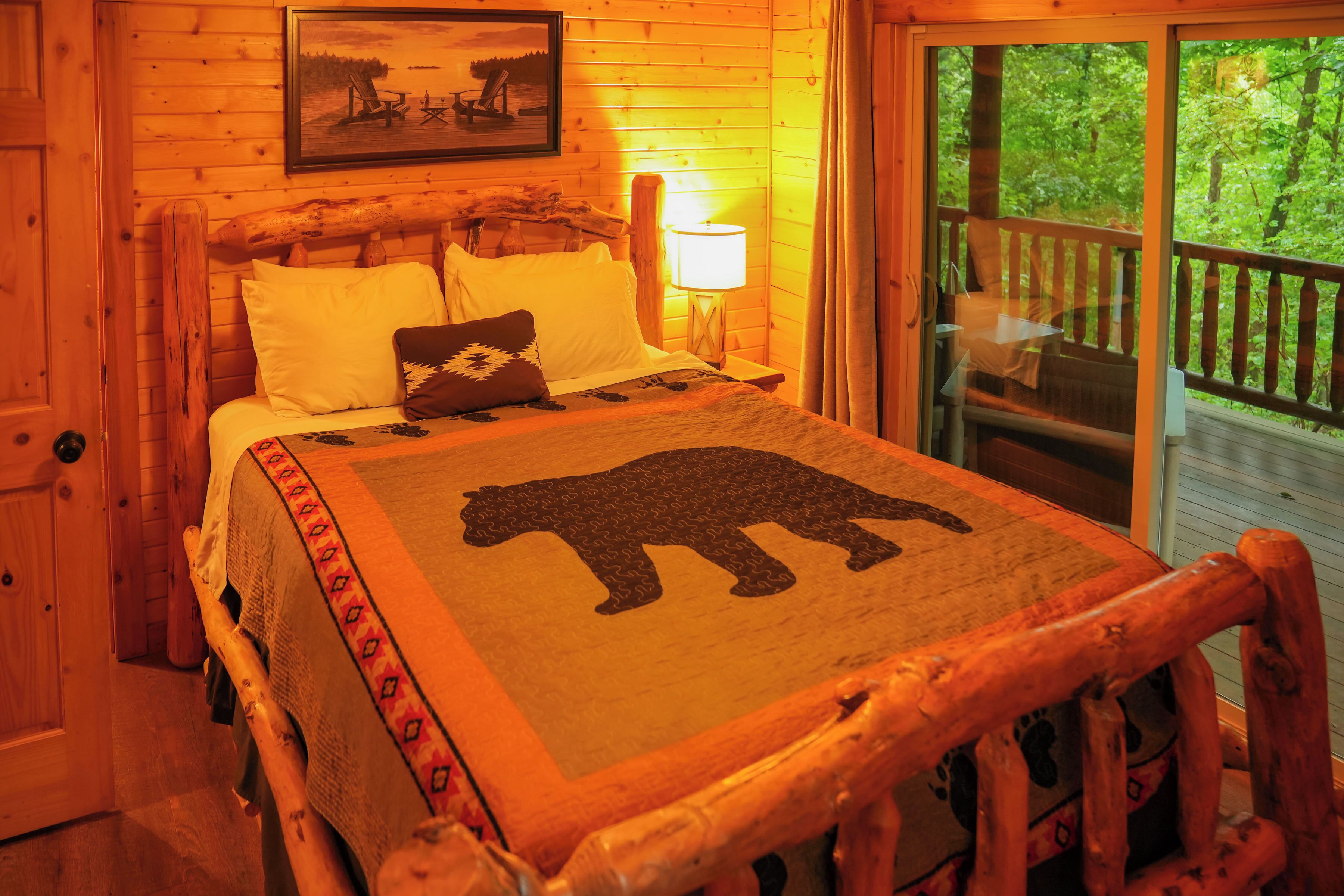 A wooden cabin room with a bed, a nightstand, a lamp, and a window with a view of trees.