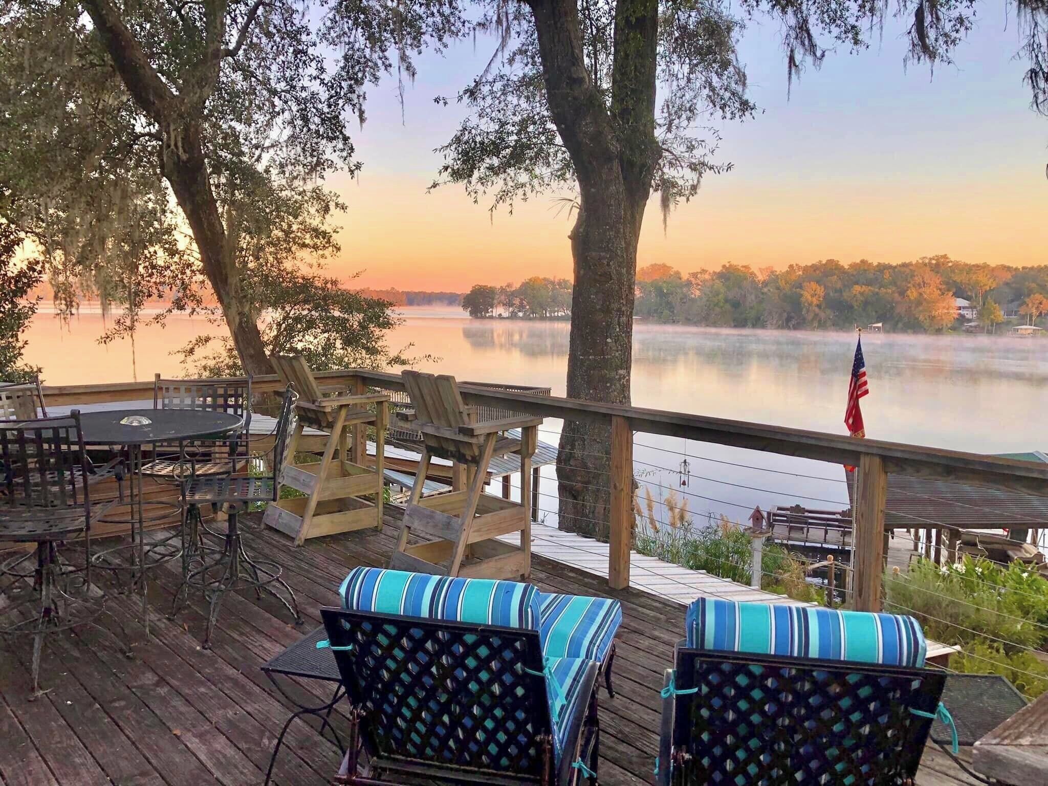 Une terrasse avec coin salon extérieur, offrant une vue sur un lac paisible, bordé d’arbres, et un ciel dégagé.
