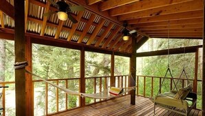 A wooden deck with a hammock and hanging chair, large windows, and ceiling fans.