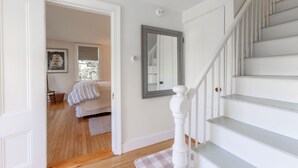 Una escalera con barandillas blancas, piso de madera y un tapete a cuadros que conduce a un dormitorio con cama y ventana.