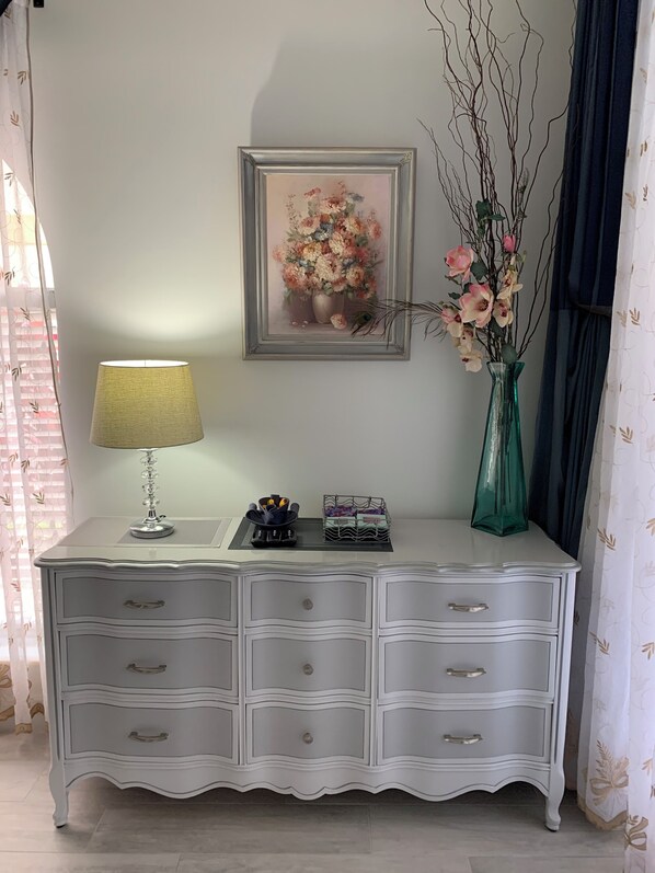 Een witte dressoir met lades, een lamp, een lijst met bloemstilleven en een vaas met bloemen.