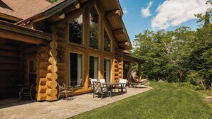 A log cabin with a patio area featuring a table and chairs, surrounded by a well-maintained lawn and trees. - ChaletsOais Luxury lakefront 5 bedroom log home with private hot tub  (Mille-Isles)