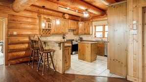 A log cabin-style kitchen with wooden cabinets, a central island, bar stools, and a built-in stove. - ChaletsOais Luxury lakefront 5 bedroom log home with private hot tub  (Mille-Isles)