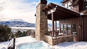 A wooden cabin with a stone chimney, a hot tub, and a view of a snow-covered mountain.