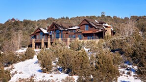 A large, multi-story lodge with a stone facade and multiple balconies, surrounded by snow-covered trees and a clear sky.