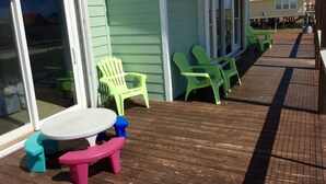 A covered outdoor area with a wooden deck, green Adirondack chairs, a small table, and a pink bench. - Family Friendly, Ft Morgan Beach House (Gulf Shores)