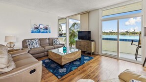 A spacious living room with a leather sofa, a glass coffee table, and a television on a wooden cabinet. A large window offers a view of a swimming pool and palm trees.