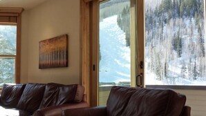 A room with a brown leather sofa, a painting on the wall, and a large window with a snowy mountain view.